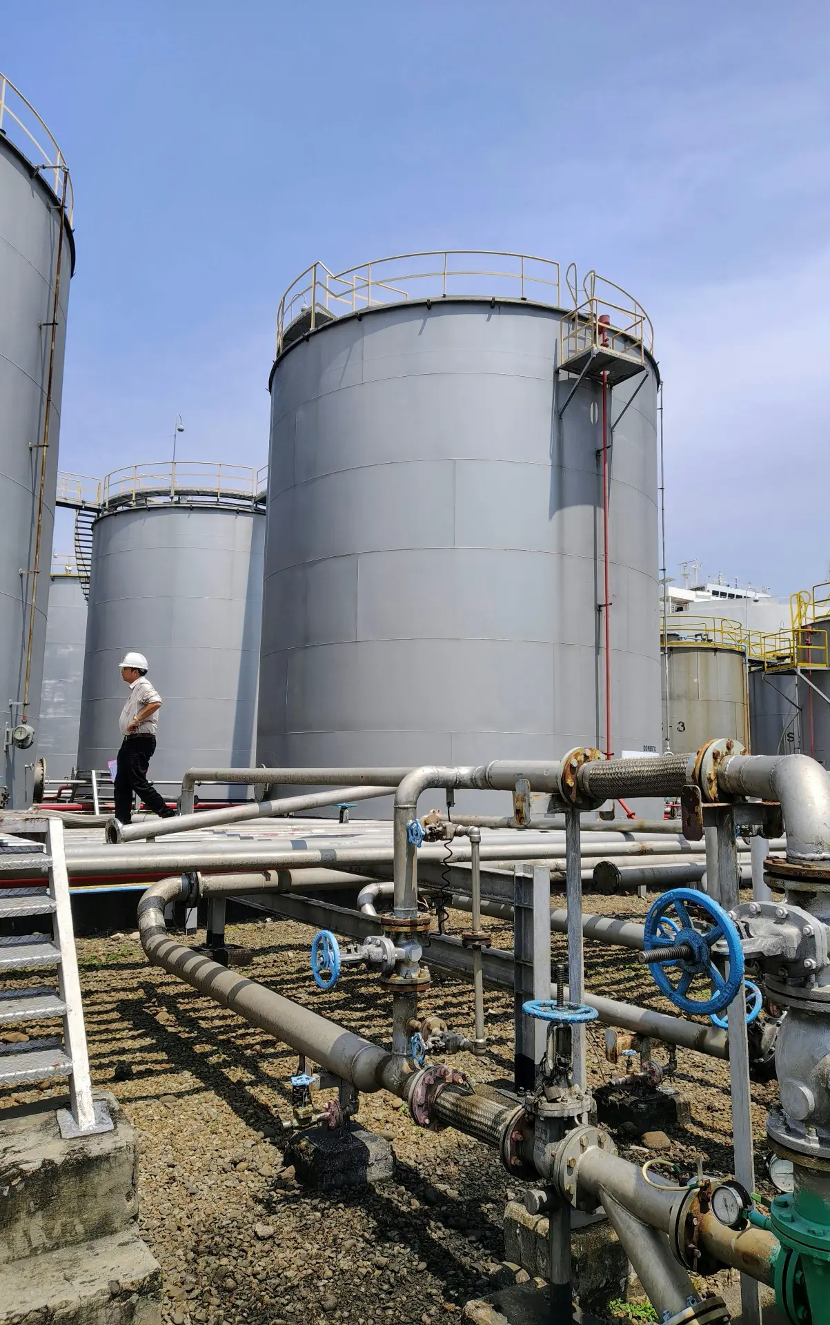 Engineer walking a tank farm past storage tanks, pipework and valve manifolds.